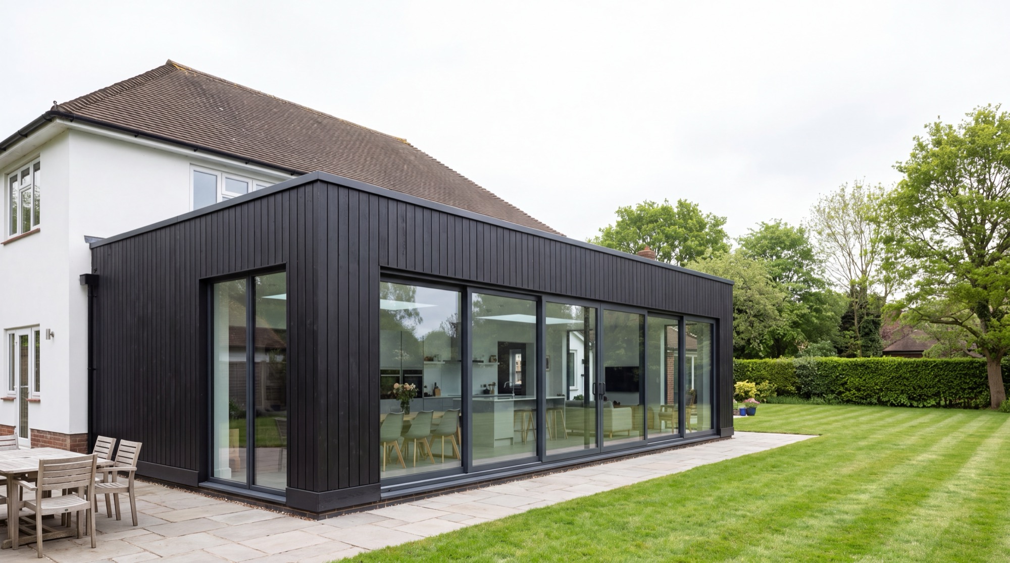 A large Nordu extension in black timber cladding across the rear of a white rendered house, glazed corner to corner along its garden elevation, with a kitchen and dining table visible inside.